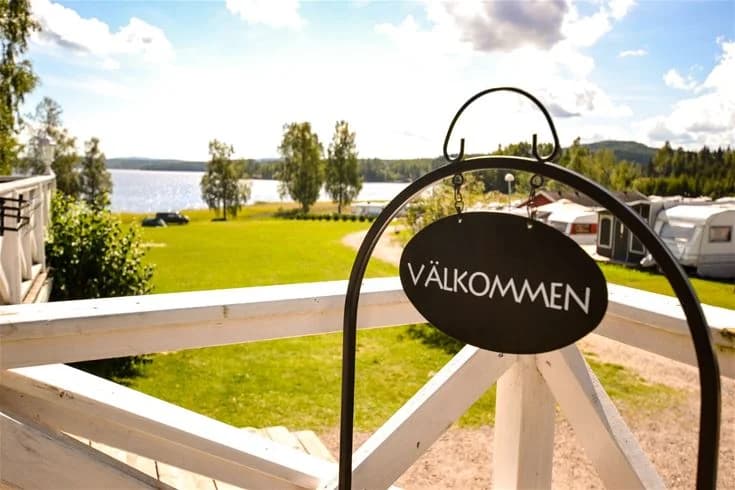 Bild av Charlottenberg camping & stugor - Haganäset med en grönskande gräsmatta, skuggade områden och landskapsdesign. I bakgrunden syns en bondgårdsliknande byggnad bland parkerade husvagnar och stugor.