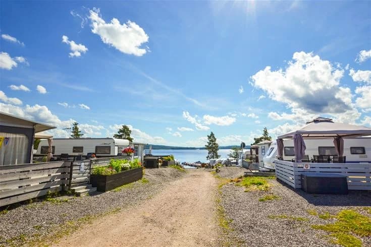En husvagn står parkerad vid strandkanten på Charlottenberg camping & stugor - Haganäset, med en vy över stranden och viken under en himmel med cumulusmoln.