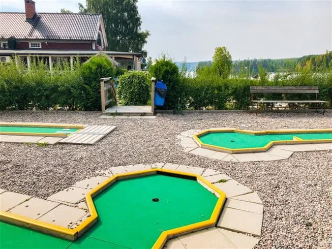 En grupp människor spelar minigolf på konstgräs i ett rekreationsområde vid Charlottenberg camping & stugor - Haganäset. I bakgrunden syns grönskande gräsmattor och träd, vilket skapar en avslappnad och naturnära atmosfär.