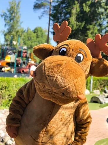 En glad älgmaskot leker i ett semesterlandskap vid Charlottenberg camping & stugor - Haganäset, omgiven av grönskande gräs och symboliserar glädje och gemenskap.
