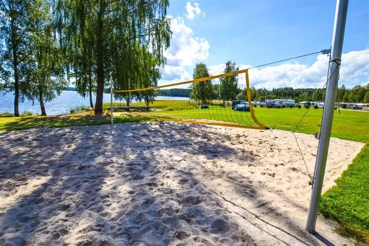 En sandstrand med ett uppsatt volleybollnät, omgiven av träd och parkmiljö på Charlottenberg camping & stugor - Haganäset.