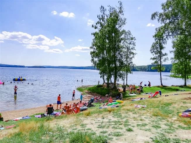 En somrig vy av Charlottenberg camping & stugor - Haganäset, med en sjö och omgivande kustlandskap. Människor njuter av fritidsaktiviteter vid strandkanten.