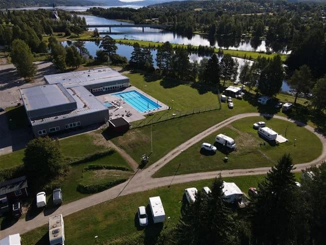 En campinganläggning med en simbassäng omgiven av grönska och tallar, belägen i en lugn rekreationsmiljö som erbjuder olika vattensporter.