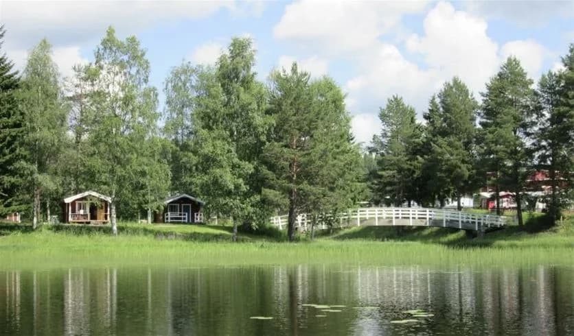 En lugn och naturskön vy över Stöde Camping med träd och grönska vid kanten av en sjö, gräsmatta i förgrunden.