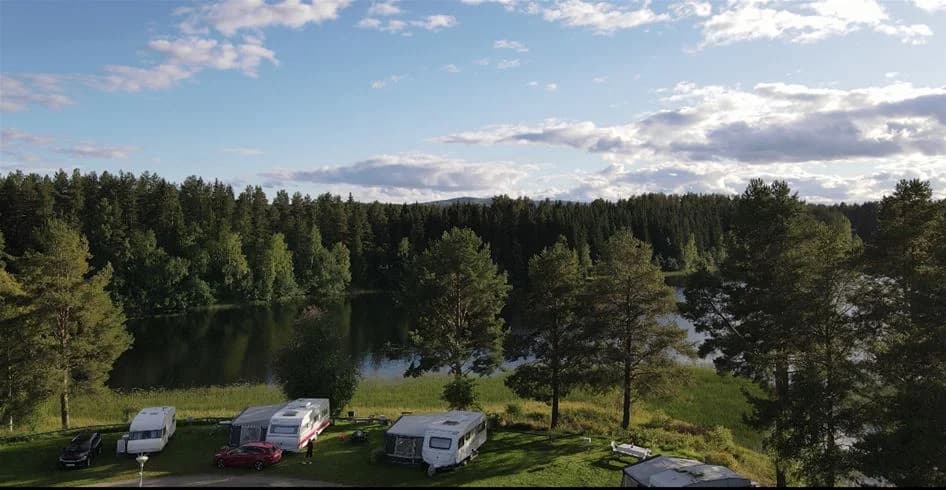 En naturskön vy över Stöde Camping med en glittrande sjö i förgrunden, omgiven av frodig grönska och höga träd som sträcker sig mot himlen.