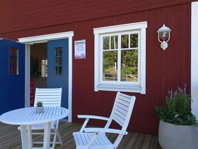 Bild på en trästuga vid Yxningens camping, med en enkel trästol utanför dörren. Stugan ger en hemtrevlig känsla med sin träpanel och lätt slitna utseende, omgiven av grönska.