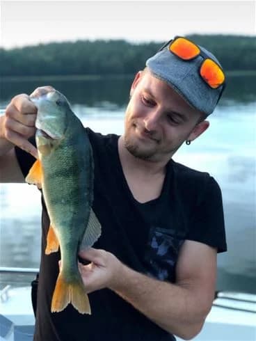 En person med keps håller upp en nyfångad abborre vid Yxningens camping, visar en lyckad fisketur.