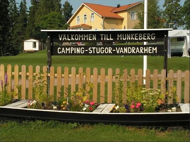 En grönskande campingplats med välklippt gräsmatta och skuggiga träd. I bakgrunden syns en mobilbostad och campingutrustning, vilket skapar en avkopplande och naturnära miljö. Perfekt för friluftsliv och rekreation.