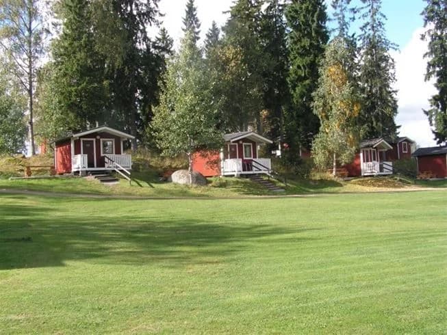 En grönskande campingplats med gräs, träd och stugor som omges av en välskött gräsmatta och trädgård. I bakgrunden syns ett stort markområde och en park. 