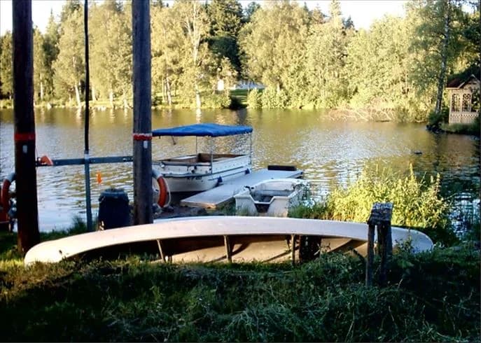 En lugn sjö med små båtar förtöjda vid en brygga, omgivna av grönskande våtmarker och träddunge, med speglingar av naturen i vattnet vid Munkebergs camping.