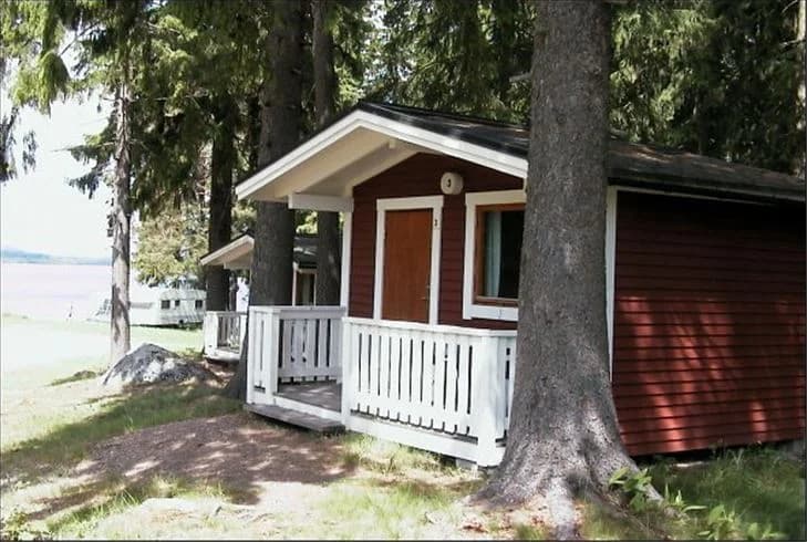 En timrad stuga omgiven av grönska på Munkebergs camping, med en veranda i skugga.