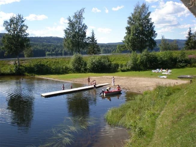 En bild av Kävsta camping visar en naturskön plats med träd och växter längs en vattenväg. En båt syns på sjön, vilket bidrar till en känsla av rekreation och avkoppling.