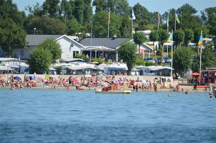 En somrig bild av Klinta Camping med människor som njuter av sol och bad vid stranden. Havet och kusten syns tydligt, vilket skapar en avslappnad och trevlig semesteratmosfär.