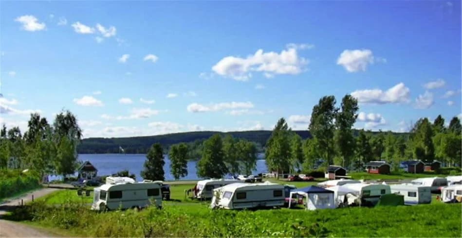 En husbil står parkerad vid strandkanten av en stilla sjö, omgiven av grönskande gräsmattor och träd. I bakgrunden syns en naturskön vy med sjön och omgivande skogar.