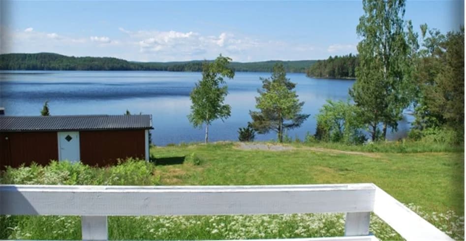 En vacker utsikt över Sölje Camping med en grön gräsmatta som leder ner till en glittrande sjö. I bakgrunden syns en liten stuga omgiven av frodig natur.