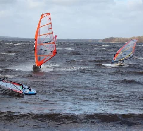En vindsurfare glider över vattnet nära Bromölla camping & vandrarhem, med segel hissat mot en klar himmel. I bakgrunden syns båtar och personer som njuter av olika vattensporter.