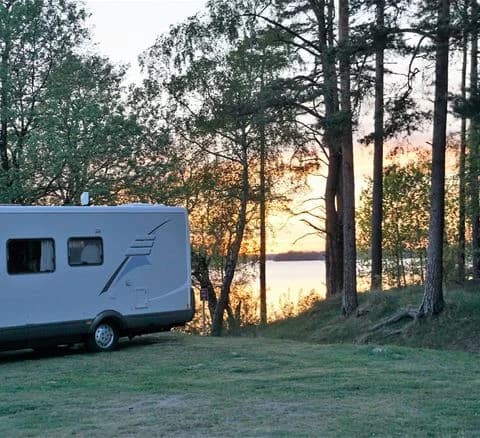 En husvagn parkerad i en skuggig del av en campingplats, omgiven av träd och grönska under klar himmel, vid Bromölla Camping & Vandrarhem.