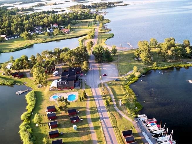 Fågelperspektiv över Dalskärs Camping vid en sjö, omgiven av grönska och små byar, med kustlinjen och kanaler i bakgrunden.