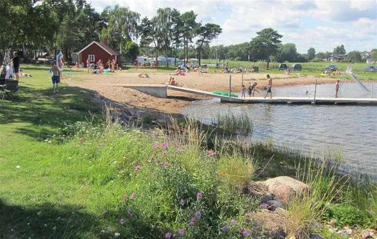 En somrig vy över Dalskärs camping med en sjö i bakgrunden. Besökare njuter av fritidsaktiviteter vid strandkanten nära en liten brygga.