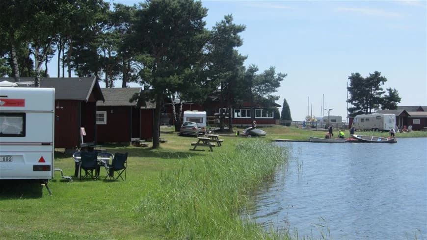 Bild av Dalskärs camping med en sjö i förgrunden där flera båtar ligger förtöjda. Grönt gräs och träd omger sjön, vilket ger en naturskön och avkopplande atmosfär.