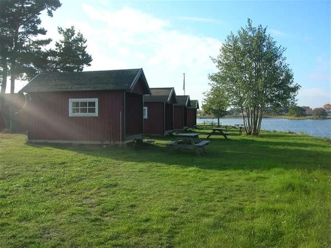 En naturskön bild från Dalskärs Camping med en vy över en lugn sjö och omkringliggande grönska. I förgrunden syns små stugor och lusthus, omslutna av välskötta gräsmattor och parkmiljö.