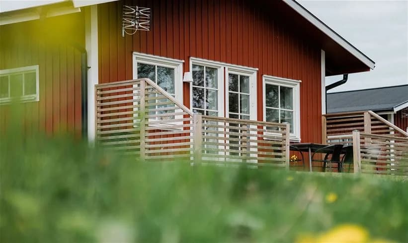 Bild av en stuga vid Mösseberg Camping med en inbjudande veranda. Stugan är gjord av trä och har en träfärgad fasad. På verandan finns utemöbler och omgivningen inkluderar en gräsmatta.
