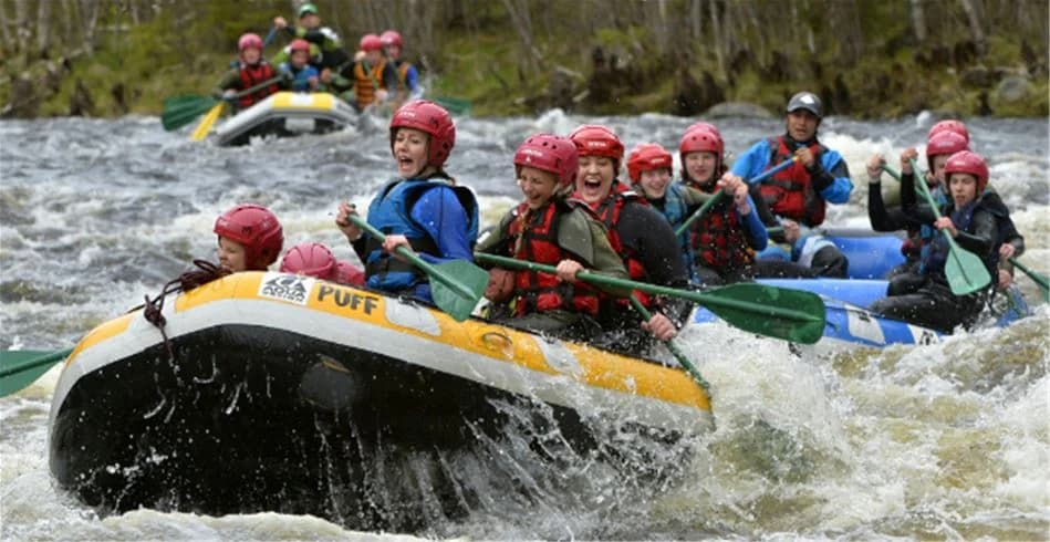 En grupp människor i skyddsutrustning paddlar i en båt nedför en fors, omgiven av frodig natur och strömmande vatten på Idre Fjäll Camping.