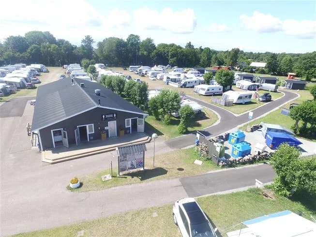 Flygbild över Sandbybadets camping som visar en campingplats med husbilar och husvagnar uppställda i ett bostadsområde. Parkeringen är synlig och omges av öppna gräsytor och små bostäder i utkanten.