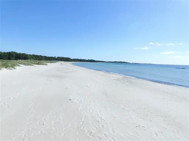 En solig dag vid Sandbybadets camping med en blå himmel och ett rofyllt hav i bakgrunden. Sandstranden sträcker sig längs kusten, vilket skapar en inbjudande miljö för besökare.