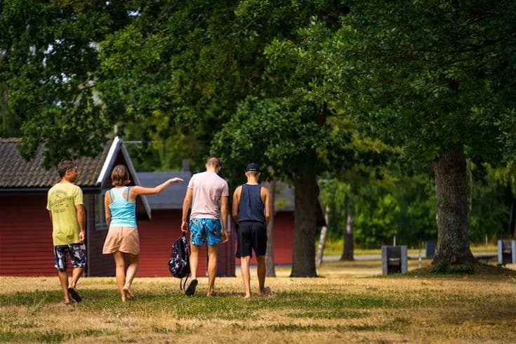 En grupp människor njuter av sommaren på Haga Park Camping & Stugor, omgivna av träd som erbjuder skugga. Solen skiner och bidrar till en avslappnad atmosfär, vilket gör det till en idealisk plats för rekreation och semester.
