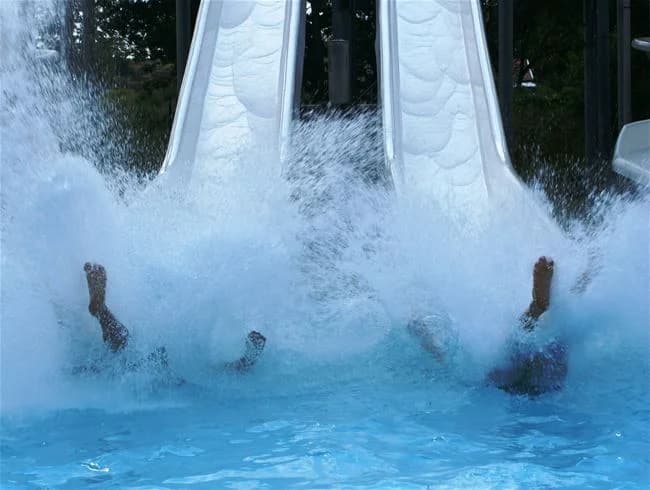 En livlig campingplats vid vattnet med människor som njuter av olika fritidsaktiviteter, inklusive en vattenpark med rutschkanor och utomhuslekutrustning, vilket bidrar till en avslappnad semesterstämning.