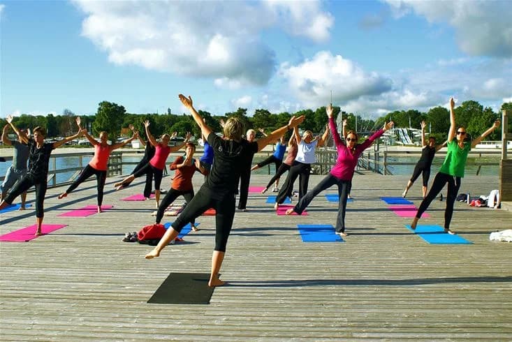 En person utför yogapositioner på en matta utomhus vid Köpingbaden Camping, omgiven av grönska och bär funktionella träningskläder. Atmosfären är avslappnad och fylld av fritidsaktiviteter.