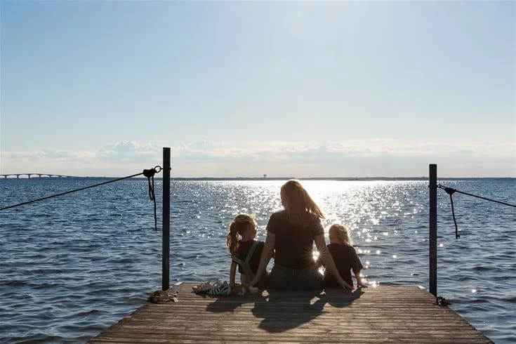 En solig sommardag vid Saxnäs camping med utsikt över en glittrande vattenyta, en lång brygga som sträcker sig ut i vattnet, och människor som njuter av avkoppling och sommaraktiviteter.