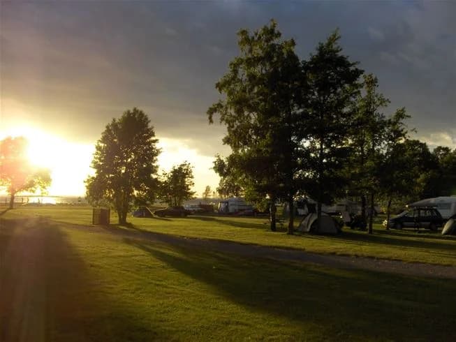 En grönskande campingplats vid saxnäs med frodig gräsmatta i förgrunden, omgiven av öppna slätter. Solens strålar skapar ett varmt ljus under skymning eller gryning, vilket framkallar en lugn och fridfull atmosfär i landskapet.