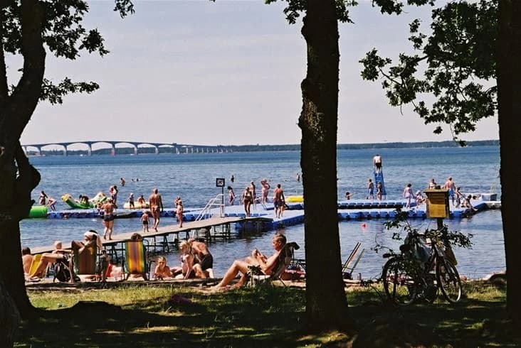 En solig dag vid kusten med besökare som kopplar av på stranden nära Saxnäs camping. Havet glittrar i bakgrunden medan människor njuter av sin semester.