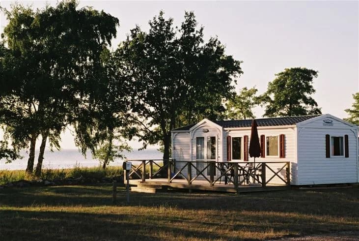 En pittoresk stuga med skuggig veranda ligger vid Saxnäs camping, omgiven av lummig grönska och några fritidshus i bakgrunden.
