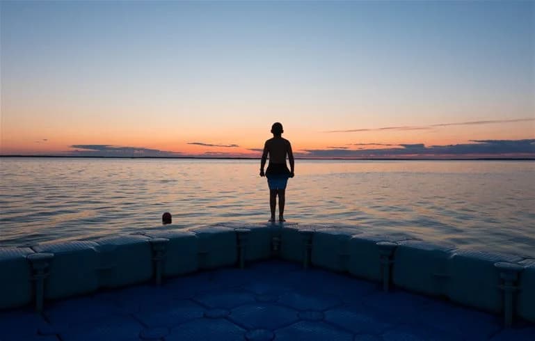 Bild av Saxnäs camping i skymningsljus, med en stilla sjö i förgrunden. Himlen domineras av mjuka moln som fångar solnedgångens varma färger, vilket skapar en reflektion i vattnet. I horisonten syns silhuetter av träd som omger campingen.