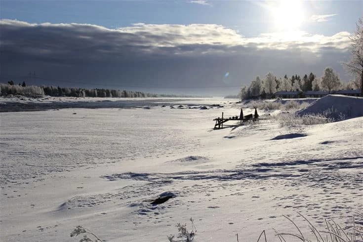 En vintrig scen vid Kukkolaforsen turist & konferens där snöklädda landskap breder ut sig under en klar himmel. Solen skapar ett mjukt ljus över de glaciärliknande formerna och sluttningarna vid forsens kanter.
