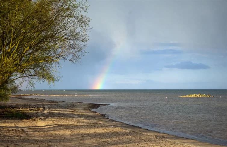 En regnbåge över en blå himmel med moln, sedd från stranden vid Stenåsa Stugor & Camping. I bakgrunden syns kusten och det öppna havet.