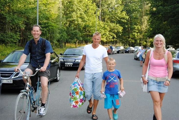 Två personer i sommarkläder cyklar längs en solig väg på Eksjö Camping & Konferens, omgivna av grönskande natur.