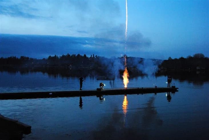 Bild av en sjö vid skymning med reflektioner i vattnet. I bakgrunden syns en eld som avger rök och värme, omgiven av en lugn och fridfull atmosfär.