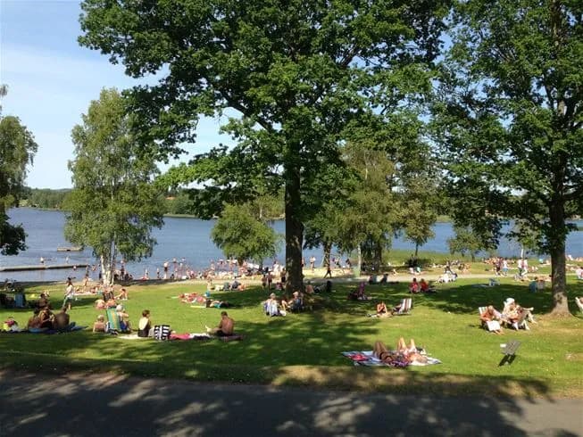 En sommardag vid Eksjö Camping & Konferens med människor som solar och har picknick vid en sjö omgiven av lummiga träd.