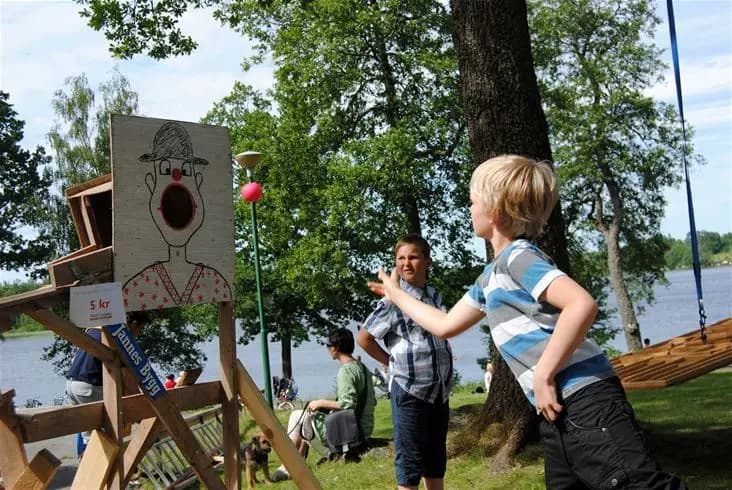 En grupp människor, bland annat barn, som leker och njuter av fritidsaktiviteter i en park vid Eksjö Camping & Konferens. Det är en solig dag som speglar en avslappnad och rolig semesterstämning.