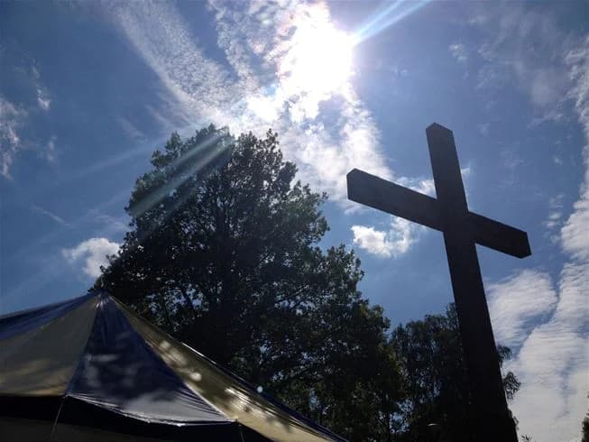 En bild av en soluppgång eller solnedgång vid Eksjö Camping & Konferens, där solen strålar genom träden. Ett träkors står i förgrunden, vilket skapar en symbolisk och rofylld stämning. Ljuset ger en varm och inbjudande atmosfär med en känsla av lugn och eftertänksamhet i naturlig omgivning.