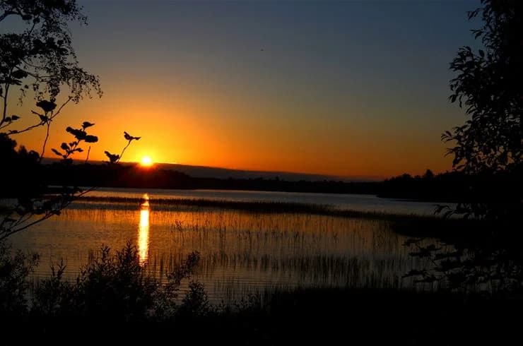 Solnedgång över en vattenyta vid Eksjö Camping & Konferens, med himlen färgad i gula och orangea toner och landskapet runt vattnet insvept i skymningsljus.