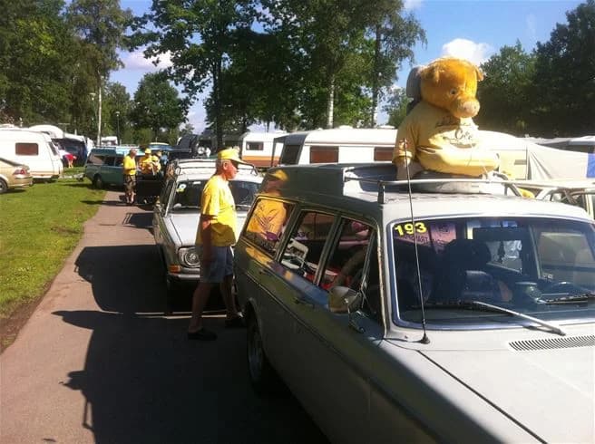 En familjebil med lastbärare på taket står parkerad vid en stugliknande byggnad på Eksjö camping & konferens. Bilens baklyktor och dörrar är synliga, och bredvid finns ett räcke som avgränsar området.