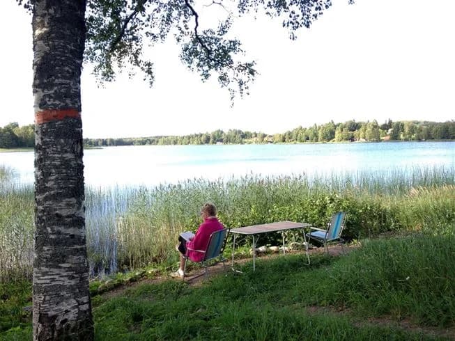 En grön och somrig campingplats vid sjön, med gräs och växtlighet i förgrunden. Utemöbler syns vid strandkanten, vilket ger en inbjudande atmosfär för besökare.