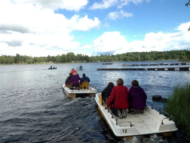 En bild som visar människor som njuter av båtliv på en sjö vid Eksjö Camping & Konferens. Flera båtar syns på den lugna vattenytan omgivna av grönskande landskap. Perfekt för utomhusaktiviteter och rekreation.