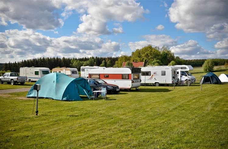 En campingplats vid Sannabadet med tält, husvagnar och en husbil på en gräsbevuxen yta omgiven av en äng och parkerad lastbil.