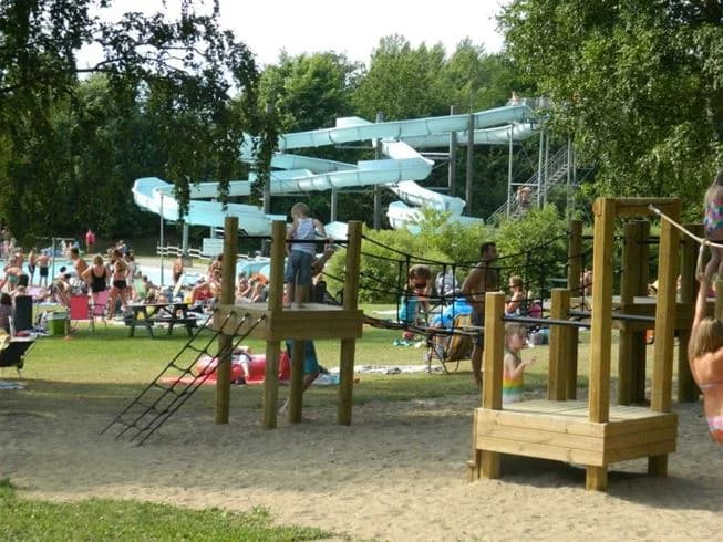 En solig dag vid Sannabadet, där människor njuter av friluftsliv och rekreation i en parkmiljö. Lekplatsutrustning syns bland träden, och besökare klädda i badkläder deltar i sommaraktiviteter och svalkar sig i det omgivande området.